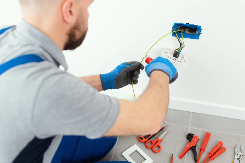 Électricien professionnel en chemise à carreaux travaillant sur installation électrique résidentielle, manipulant fils et connexions dans boîtier mural blanc